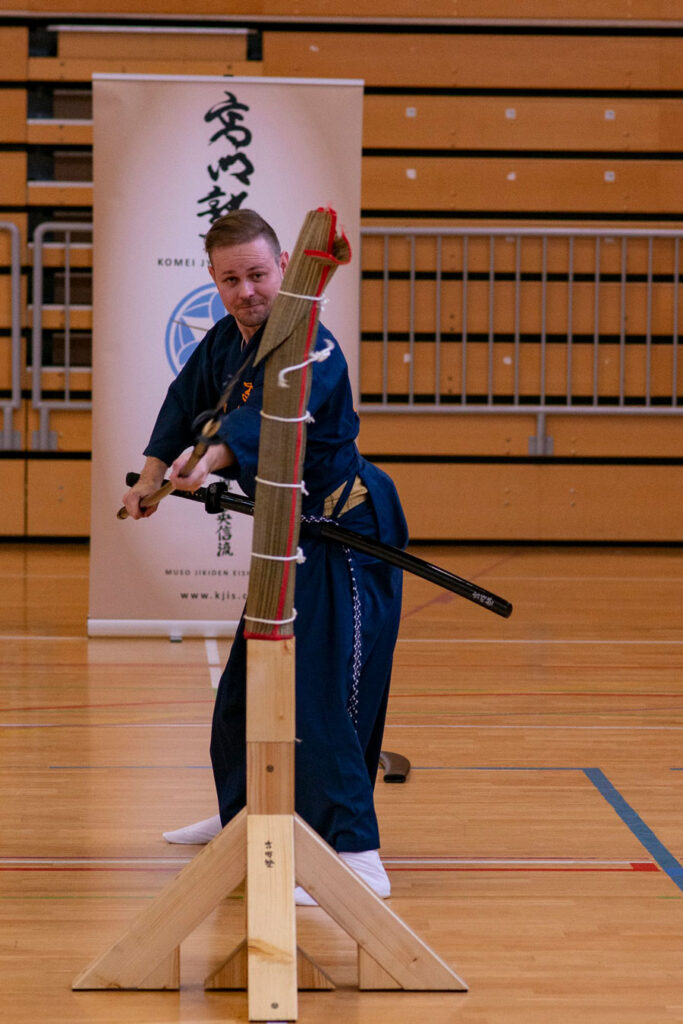 Sebastijan Sekol, Hauptlehrer von Schwertkunst Graz, in blauer Uniform beim Tameshigiri-Training in der Turnhalle – hält den Tameshigiri-Stapel vor dem Schnitt